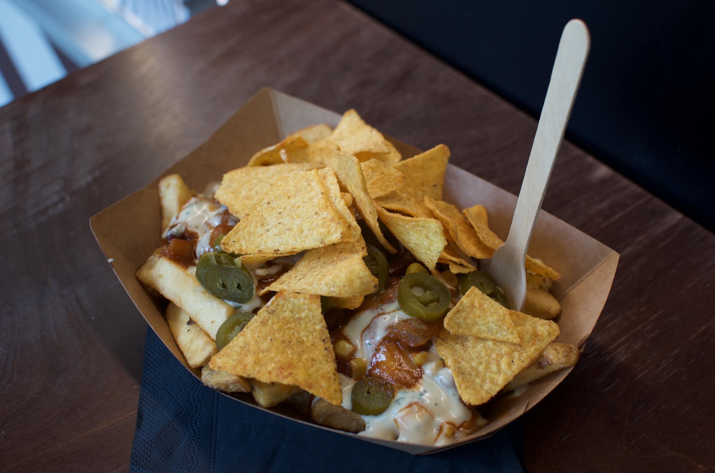Loaded fries topped with nacho chips, jalapeño slices, cheese sauce, and salsa in a brown paper tray with a wooden fork.