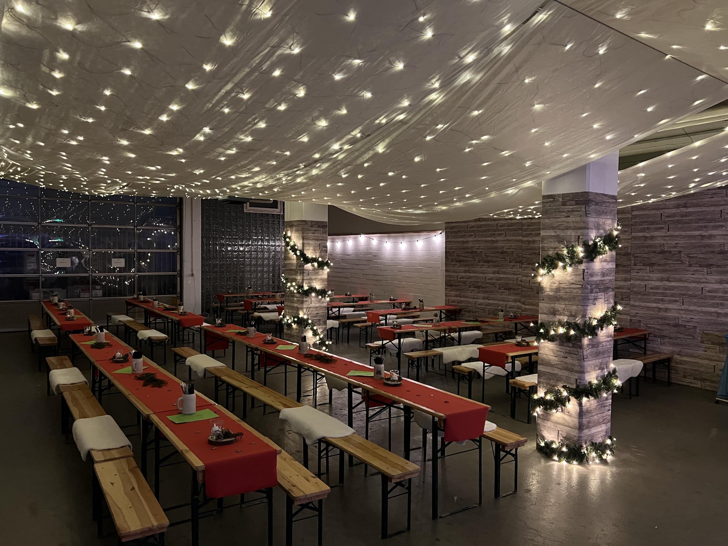 Indoor dining area with long wooden tables and benches, decorated with red table runners, white cushions, and string lights wrapped around columns and ceiling.