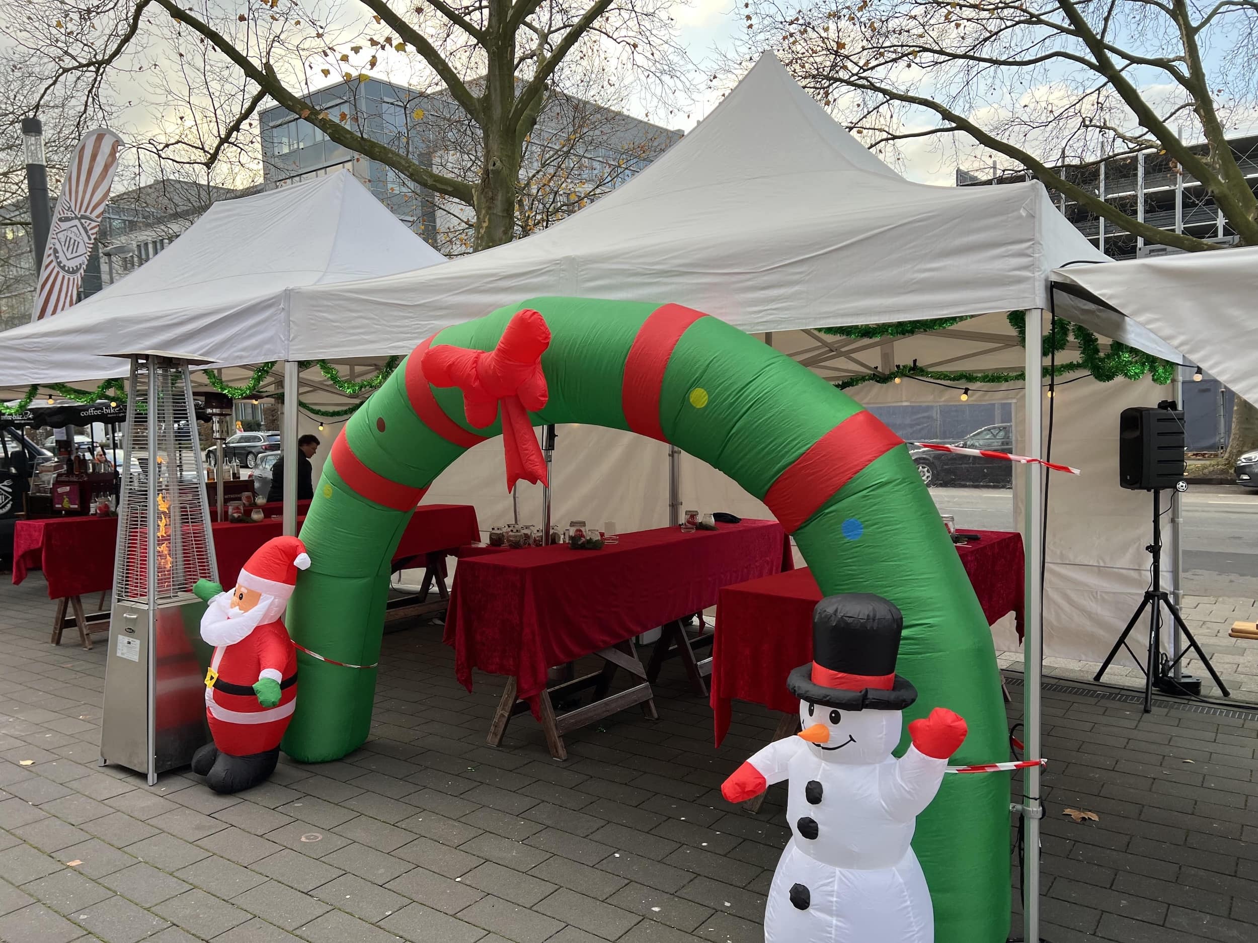 Outdoor market stalls with red tablecloths decorated with a green inflatable arch featuring a Santa figure on one side and a snowman on the other.