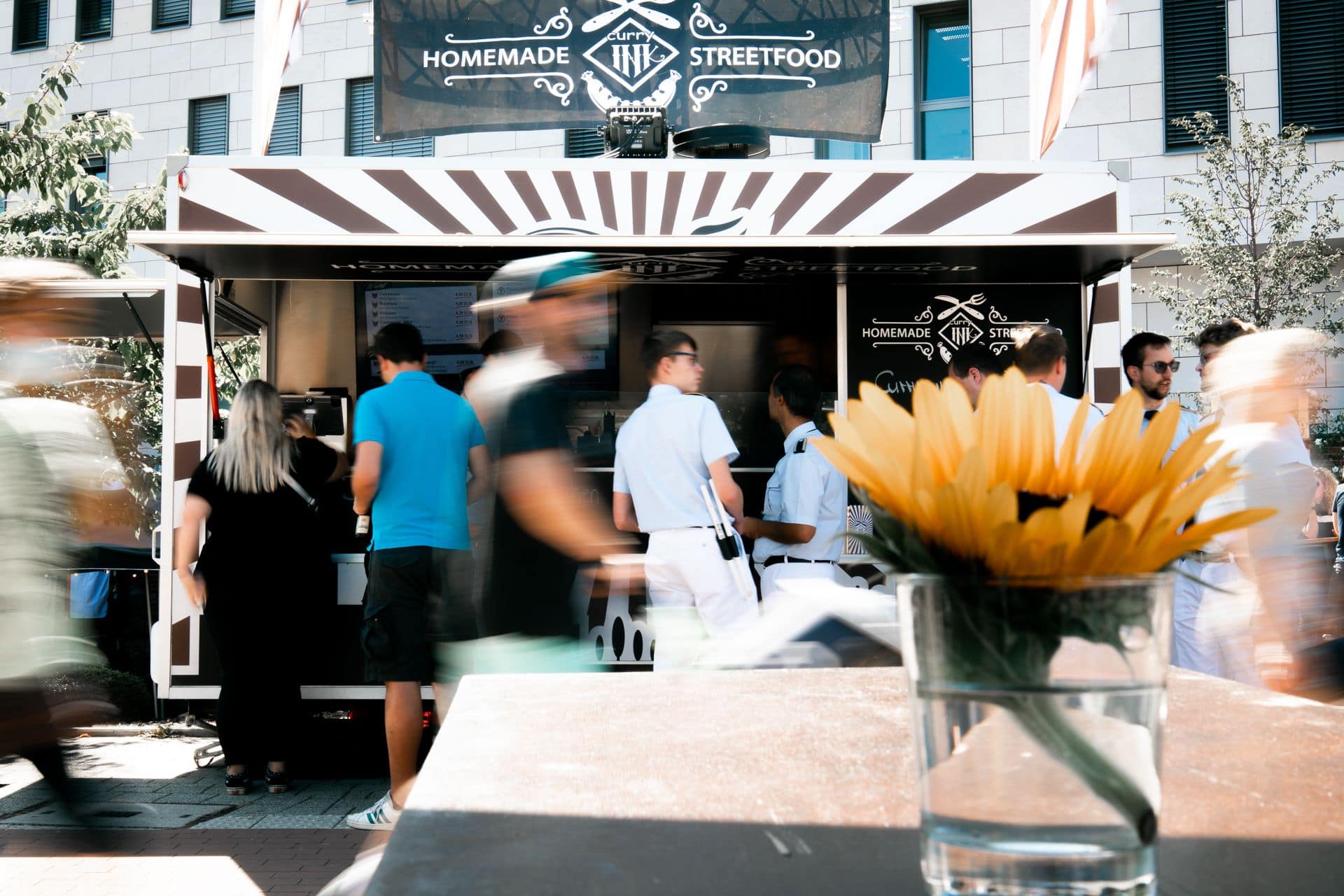Sunflower in a glass vase on a table in the foreground with a busy homemade street food truck and people in the background.